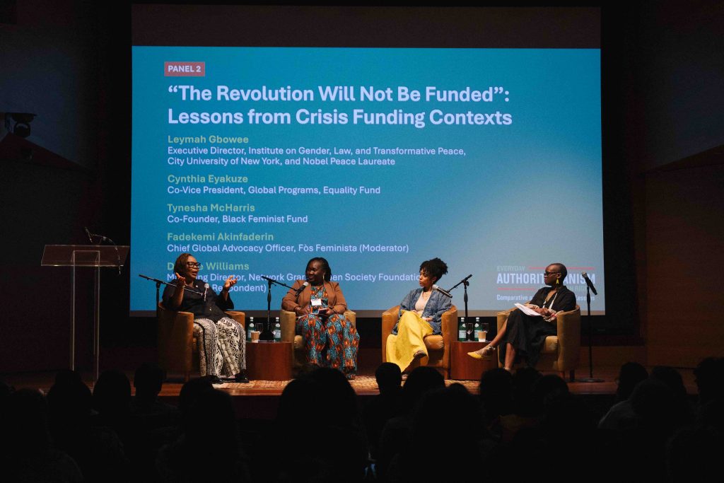 A panel of five individuals sits on a stage under a large screen displaying, “The Revolution Will Not Be Funded”: Lessons from Crisis Funding Contexts.”
