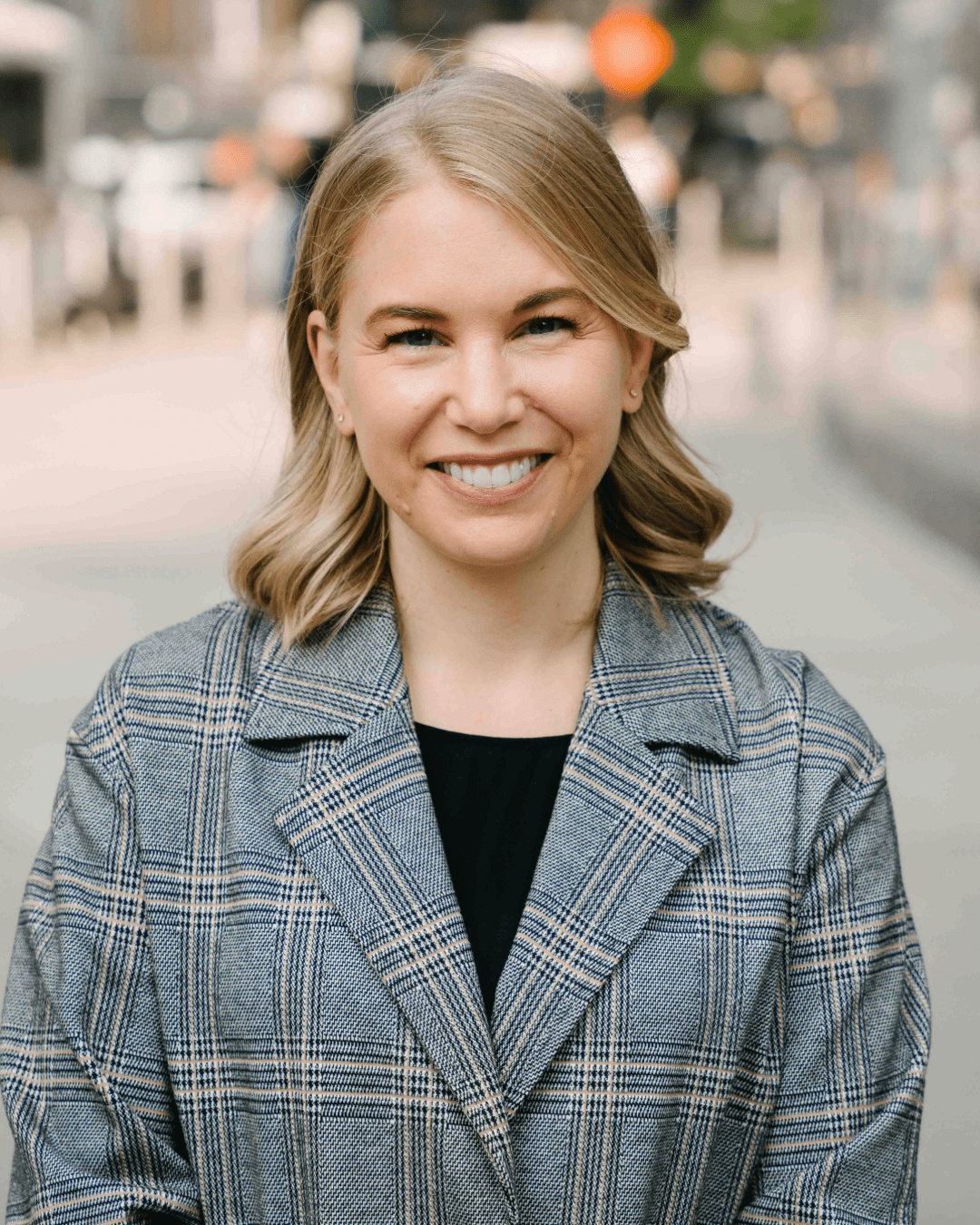 A smiling woman with blonde hair styled in loose waves, wearing a gray plaid blazer over a black top, standing outdoors on a city street.