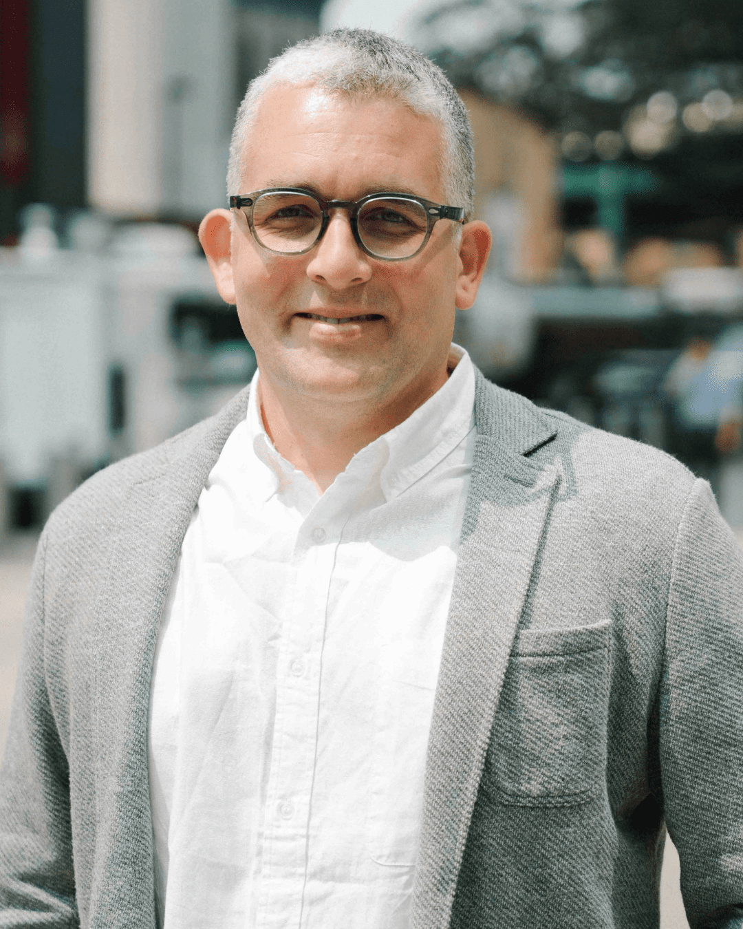 A man with short gray hair and round black glasses is smiling outdoors. He is wearing a white button-up shirt under a light gray textured blazer. The background is slightly blurred but shows an urban setting with buildings and trees.