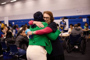 Janet Calvo hugs a participant at the Citizenship Now! event space. One wears a green blazer, and the other wears a black CUNY Law T-shirt. Tables filled with attendees and volunteers are in the background.