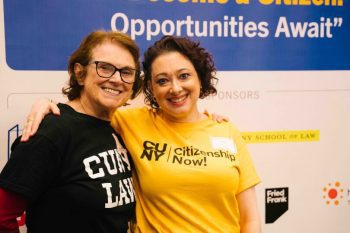 Janet Calvo and Tamara stand and pose together in front of a Citizenship Now! event backdrop. Janet wears a black CUNY Law T-shirt, and Tamara wears a yellow Citizenship Now! T-shirt. Both are smiling.