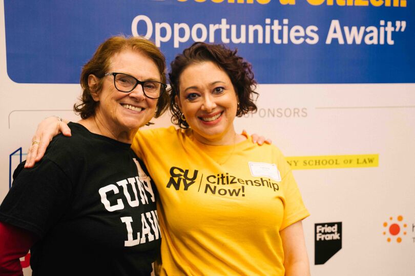 Janet Calvo and Tamara stand and pose together in front of a Citizenship Now! event backdrop. Janet wears a black CUNY Law T-shirt, and Tamara wears a yellow Citizenship Now! T-shirt. Both are smiling.