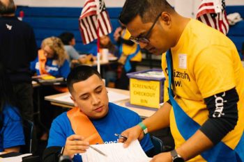 Two Citizenship Now! volunteers review paperwork together. One is seated wearing a blue T-shirt and orange sash, while the other, standing and wearing a yellow T-shirt, points to a document.