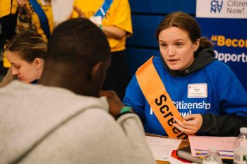 A Citizenship Now! volunteer wearing a blue T-shirt and orange “Screening” sash talks with an attendee seated across from her. Volunteers in yellow shirts and event signage appear in the background.