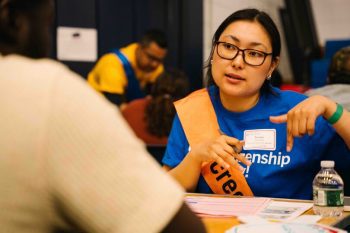 A volunteer in a blue Citizenship Now! T-shirt and orange sash speaks with an attendee, gesturing with her hands as she explains a form. Tables with other volunteers and attendees are visible behind her.
