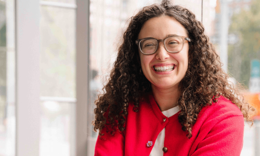 A woman with curly brown hair and glasses is smiling brightly while sitting by a large window. She is wearing a red cardigan over a white top and appears relaxed and happy. Natural light fills the space, and an urban background is visible through the window.