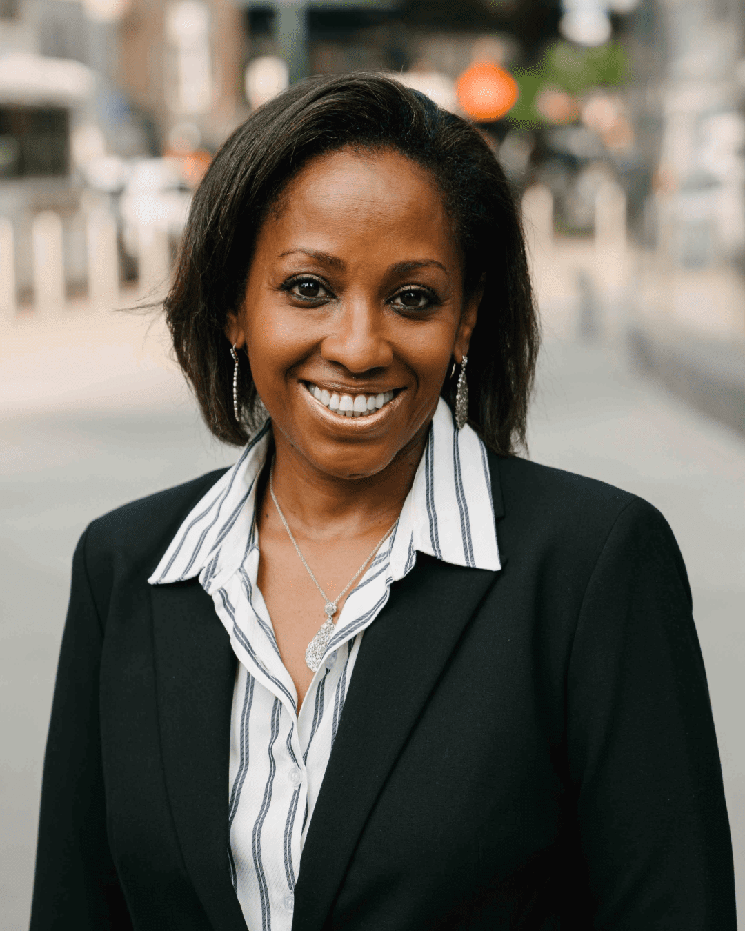 A smiling woman with straight, shoulder-length dark hair, wearing a black blazer over a white shirt with vertical black stripes, and a silver necklace, standing outdoors on a city sidewalk.