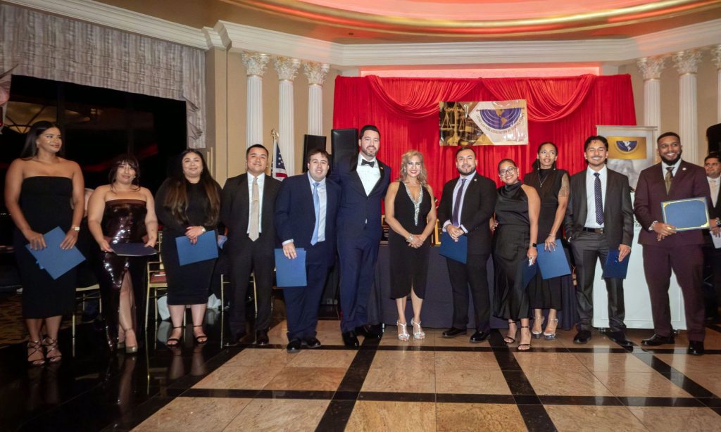 A group of law students stand side by side holding scholarship certificates at an awards ceremony hosted by the Latino Lawyers Association of Queens County. Behind them, President Robert A. Miklos, Esq., and Scholarship Committee Chair Hon. Karina E. Alomar stand beneath red drapery and the association’s banner. The event celebrates the 2025 Salvador Cheda Jr., Esq. Bar Study Scholarship recipients, marking the organization’s largest scholarship distribution to date.