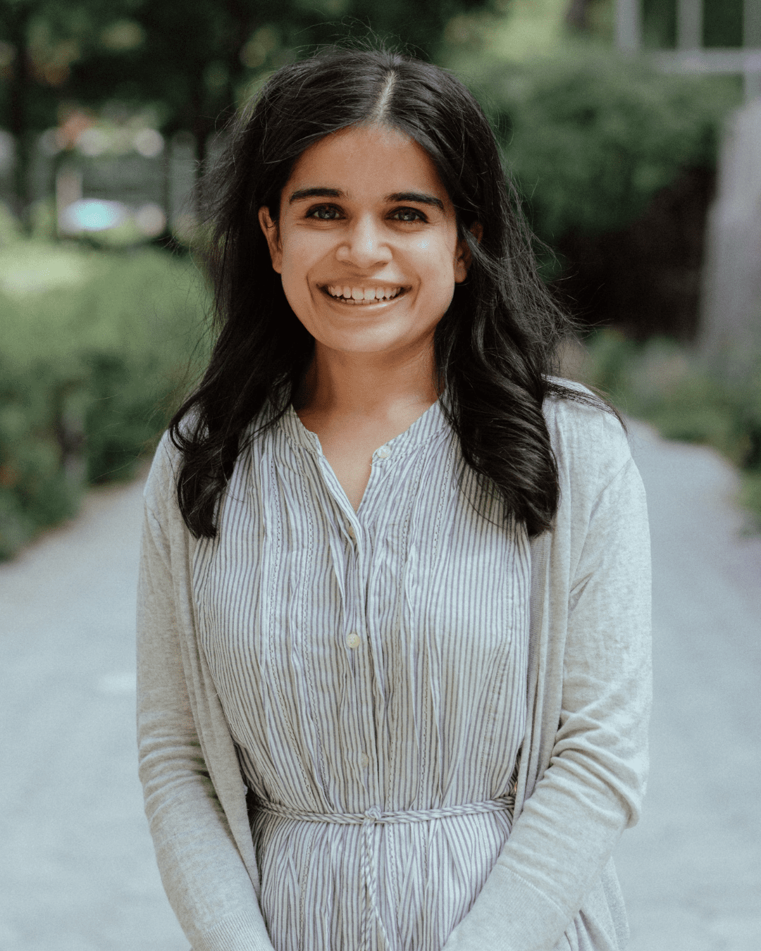 A smiling woman with long, dark, straight hair and glasses, wearing a light long sleeve dress, standing outdoors in a city park.