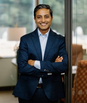 A confident man in a blue suit stands in an open doorway smiling with his arms crossed.