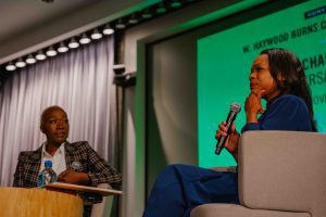 Close-up of two speakers seated in conversation; one speaks into a microphone while the other listens with a thoughtful expression.