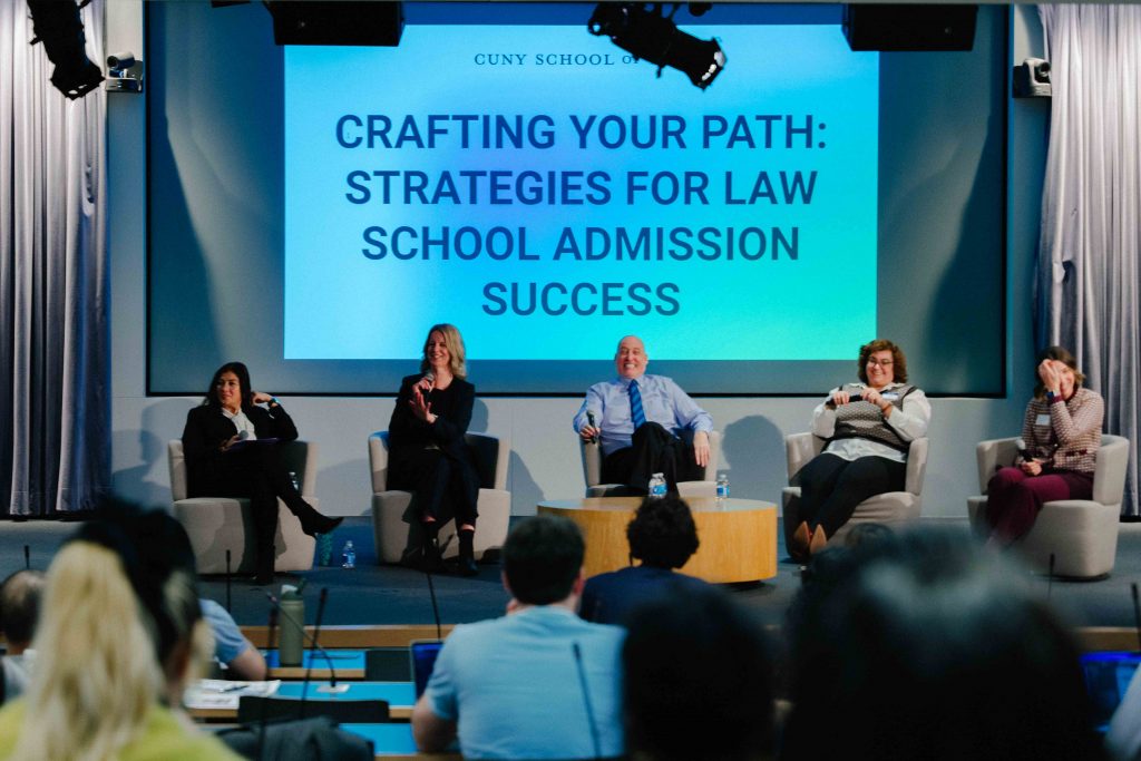 A panel of speakers sits on stage beneath a large screen that reads, “Crafting Your Path: Strategies for Law School Admission Success,” while an audience listens in the foreground.