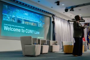 2026-01-16_Pre_Law_Conference_2nd_Annual (2 of 33) A speaker addresses the audience from the stage at CUNY School of Law, standing beside empty panel chairs. A large screen behind her reads, “Welcome to CUNY Law.”