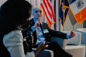2026-01-16_Pre_Law_Conference_2nd_Annual (31 of 33) A panelist speaks into a handheld microphone while seated on stage, gesturing during a discussion with U.S., New York State, and New York City flags visible behind him.