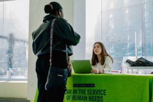 2026-01-16_Pre_Law_Conference_2nd_Annual (4 of 33) A conference registration table at CUNY School of Law where a staff member sits behind a laptop and speaks with an attendee standing in front of the table.