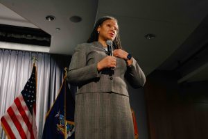 2026-01-16_Pre_Law_Conference_2nd_Annual (7 of 33) A speaker stands on stage holding a microphone and addressing the audience, with U.S. and New York State flags positioned behind her.