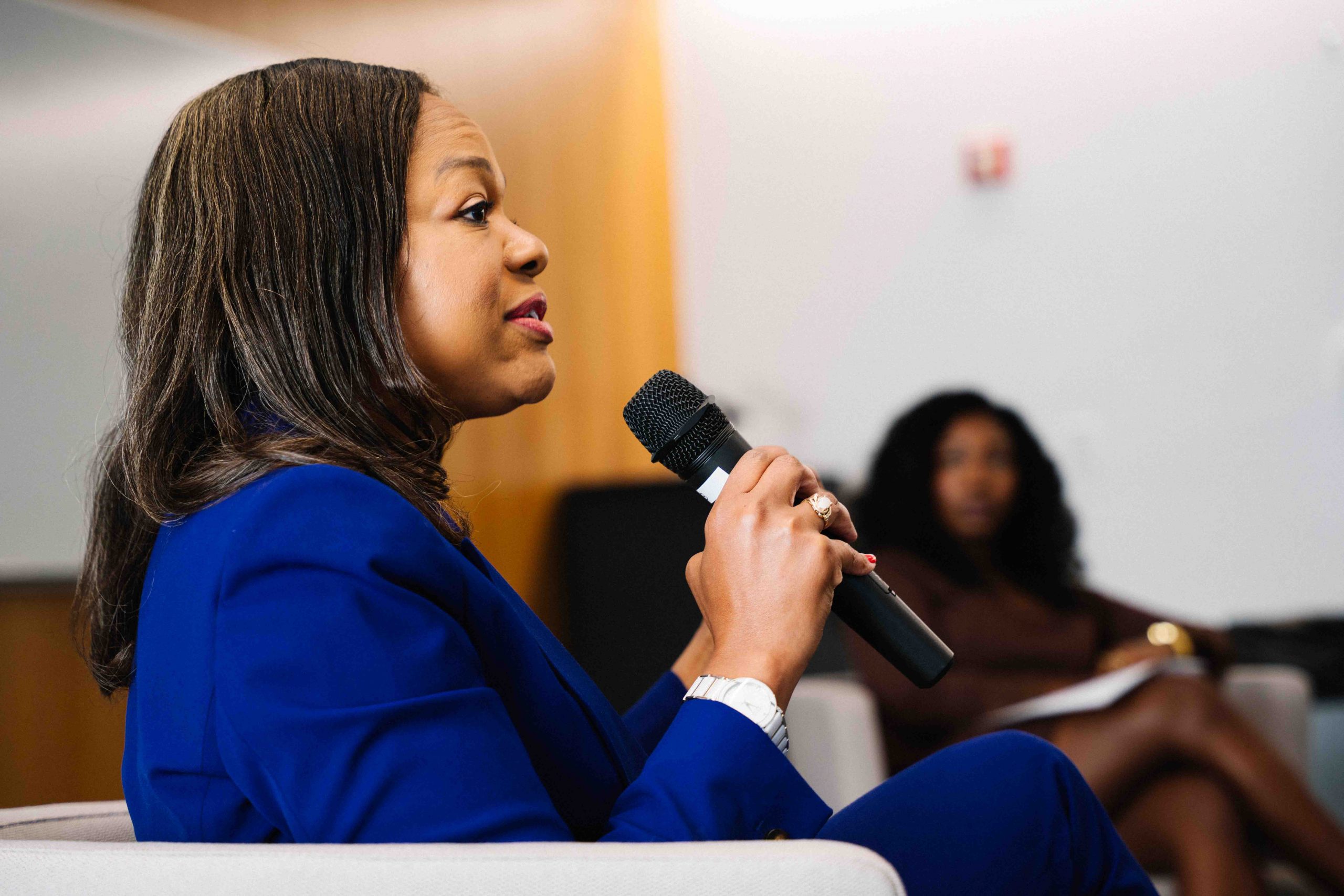 rofile view of the woman in blue speaking into a microphone, showing her from the side as she addresses the audience.