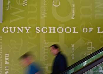 People move down the escalators in front of a wall reading "Welcome" to CUNY Law in many languages