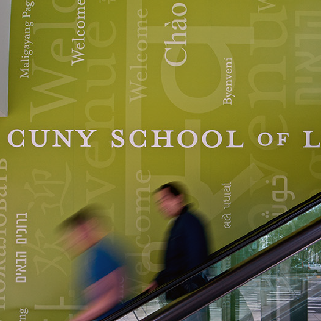 People move down the escalators in front of a wall reading "Welcome" to CUNY Law in many languages