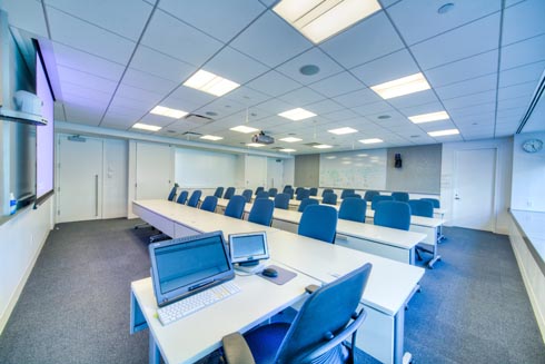 Modern classroom with digital board, white board and several rows of tables with approximately 8 seats per row for students