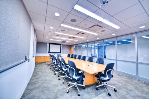 A conference room with a glass wall to the hall and a very large table in the center with at least 12 office chairs and the AV technology necessary for presentations and virtual meetings