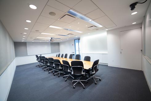 A conference room with a very large table in the center with at least 12 office chairs and the AV technology necessary for presentations and virtual meetings