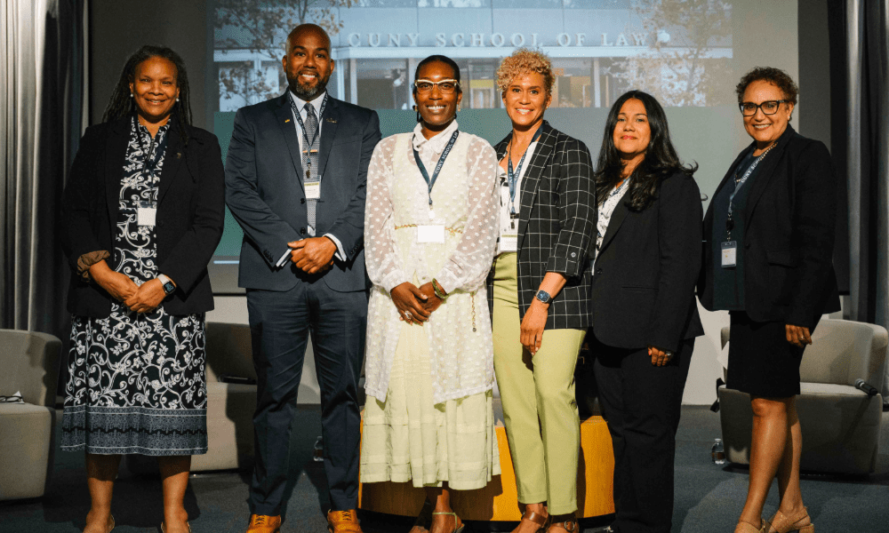 Six professionally dressed individuals stand side by side, smiling at the camera during an alumni panel event at a law school. A backdrop shows the words 'School of Law.' The group consists of four women and two men, all wearing conference badges