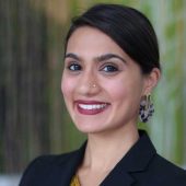 A woman in a black blazer smiles warmly, wearing bold earrings and a nose stud. The blurred green background adds a professional and approachable tone.