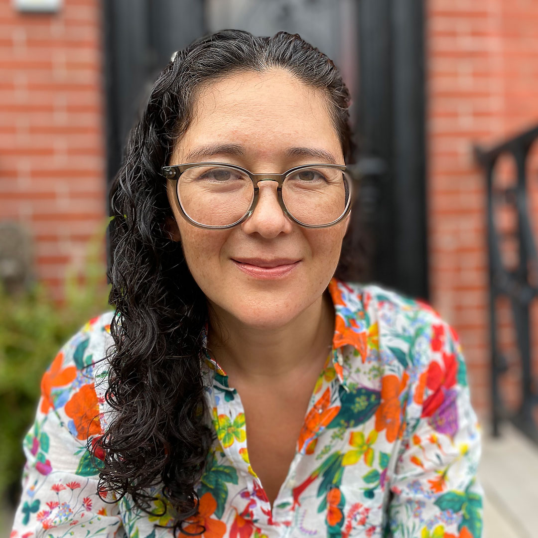 Missy sits on the front porch of a home in a floral top and glasses, smiling.