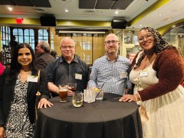 4 people at the alumni happy hour in at Busboys and Poets in DC, posed around a hightop table covered in a black tablecloth. There are wine glasses on the table. Individuals are facing the camera and smiling