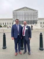 Two adults stand in front of the U.S. Supreme Court. The exterior of the Court is partially obscured by scaffolding.