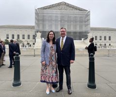 Two adults stand in front of the U.S. Supreme Court. The exterior of the Court is partially obscured by scaffolding.