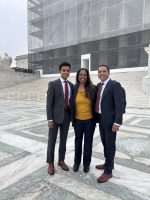 Three people stand in front of the U.S. Supreme Court. The exterior of the Court is partially obscured by scaffolding.