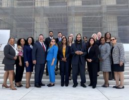 A group of 20+ people in business attire stand in front of the U.S. Supreme Court. The exterior of the Court is partially obscured by scaffolding.