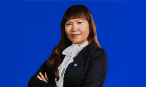 A confident woman in a black suit with a white blouse stands against a solid blue background, arms crossed, conveying professionalism and determination.