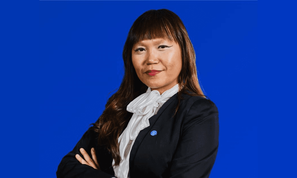 A confident woman in a black suit with a white blouse stands against a solid blue background, arms crossed, conveying professionalism and determination.