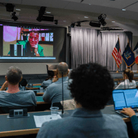 Audience members in a lecture hall watch a large screen displaying a person speaking. U.S. flags are visible; the atmosphere is focused and attentive.