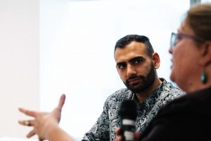 A man in a patterned shirt listens intently during a discussion, while a woman with glasses speaks into a microphone, gesturing with her hand.
