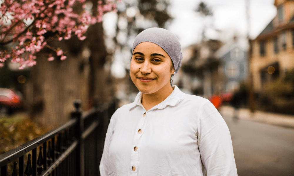 Woman in white shirt and gray headscarf standing outside on a neighborhood sidewalk and smiling.