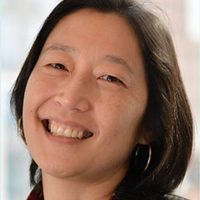Faculty_Donna-Lee_Headshot Smiling woman with short black hair, wearing hoop earrings and a dark blazer over a patterned blouse. The background is softly blurred, conveying warmth.