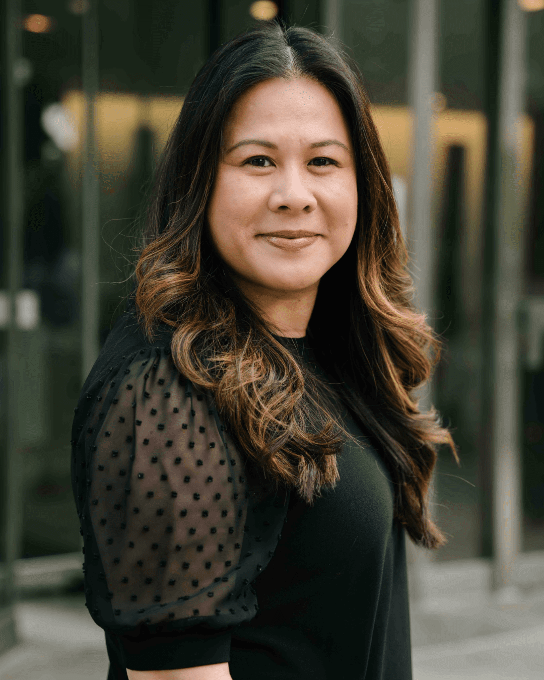 Portrait of a woman with long wavy dark hair, wearing a black top with sheer dotted sleeves, standing outdoors in front of a modern glass building
