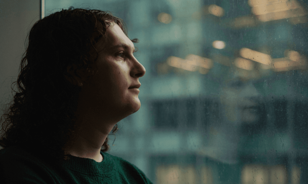 Person with curly hair gazes thoughtfully out a rain-speckled window, reflecting an urban building with a blurred silhouette. Contemplative mood.