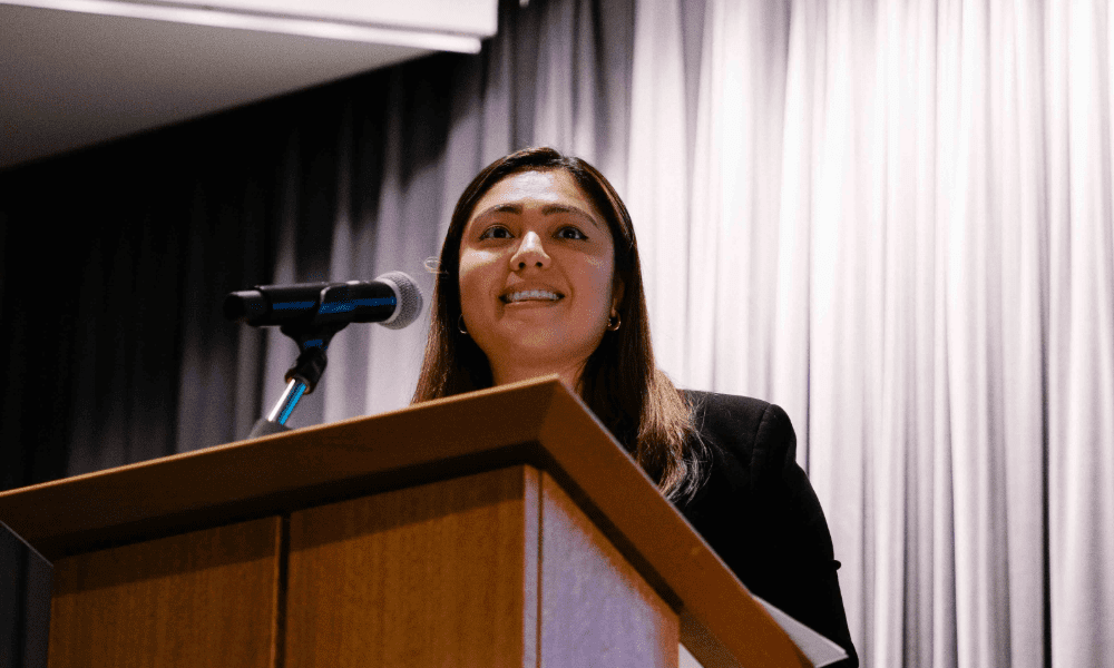 A woman stands at a podium with a microphone, speaking confidently. She is in front of a gray curtain, conveying a professional atmosphere.