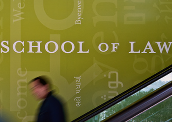 People move down the escalators in front of a wall reading "Welcome" to CUNY Law in many languages